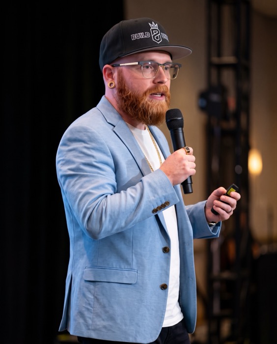 Jesse Wood speaking with a microphone in a Build Until hat and sky blue blazer